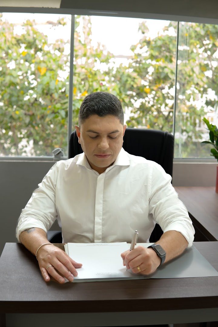 Dr. Felipe Paiva escrevendo à mão em uma folha sobre a mesa do consultório, com uma árvore na janela atrás